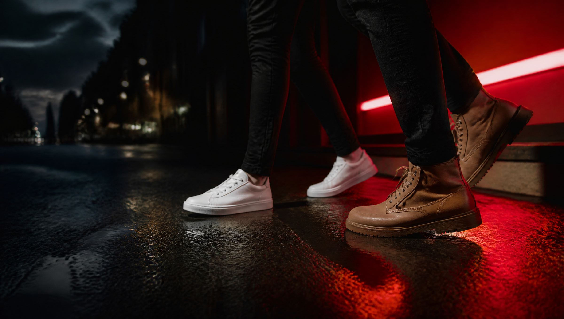 Two pairs of shoes, white sneakers and brown boots, on a wet street lit by red neon at night